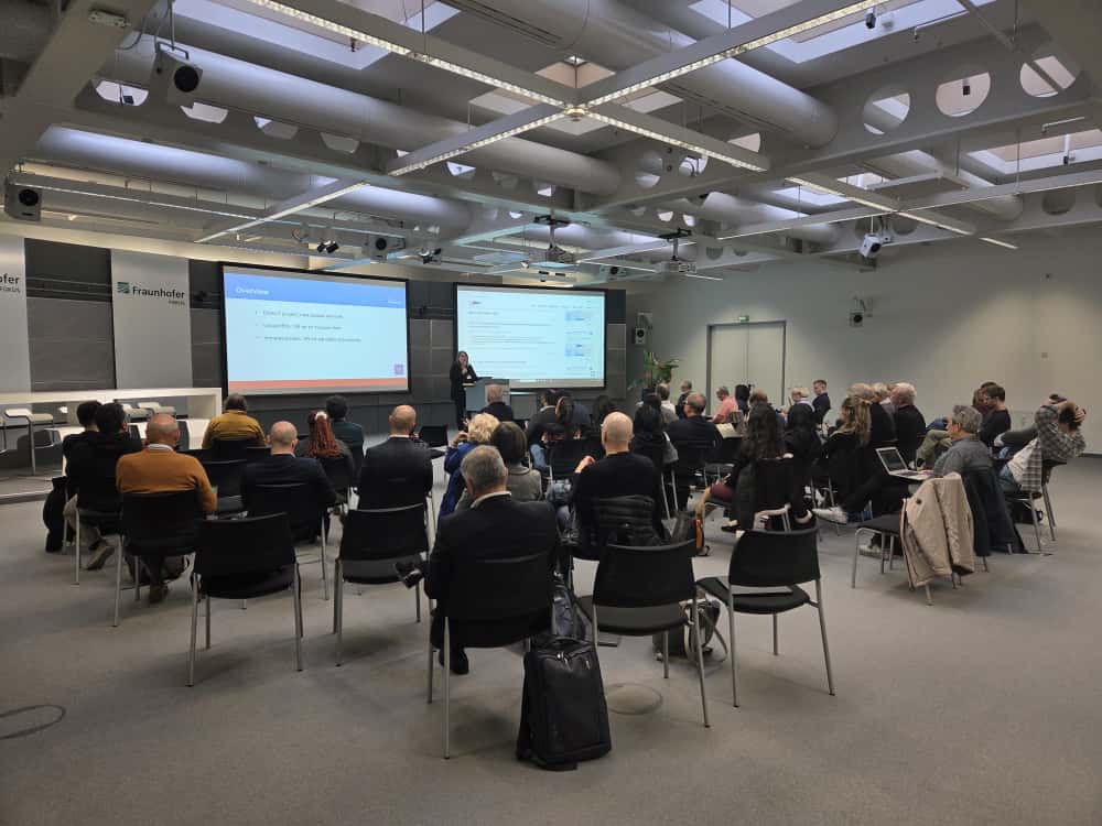 Un grup de persones assistint al NEM Summit en una sala de conferències moderna del centre Fraunhofer. Anna Matamala parla davant dues pantalles grans que mostren diapositives sobre la Xarxa AccessCat