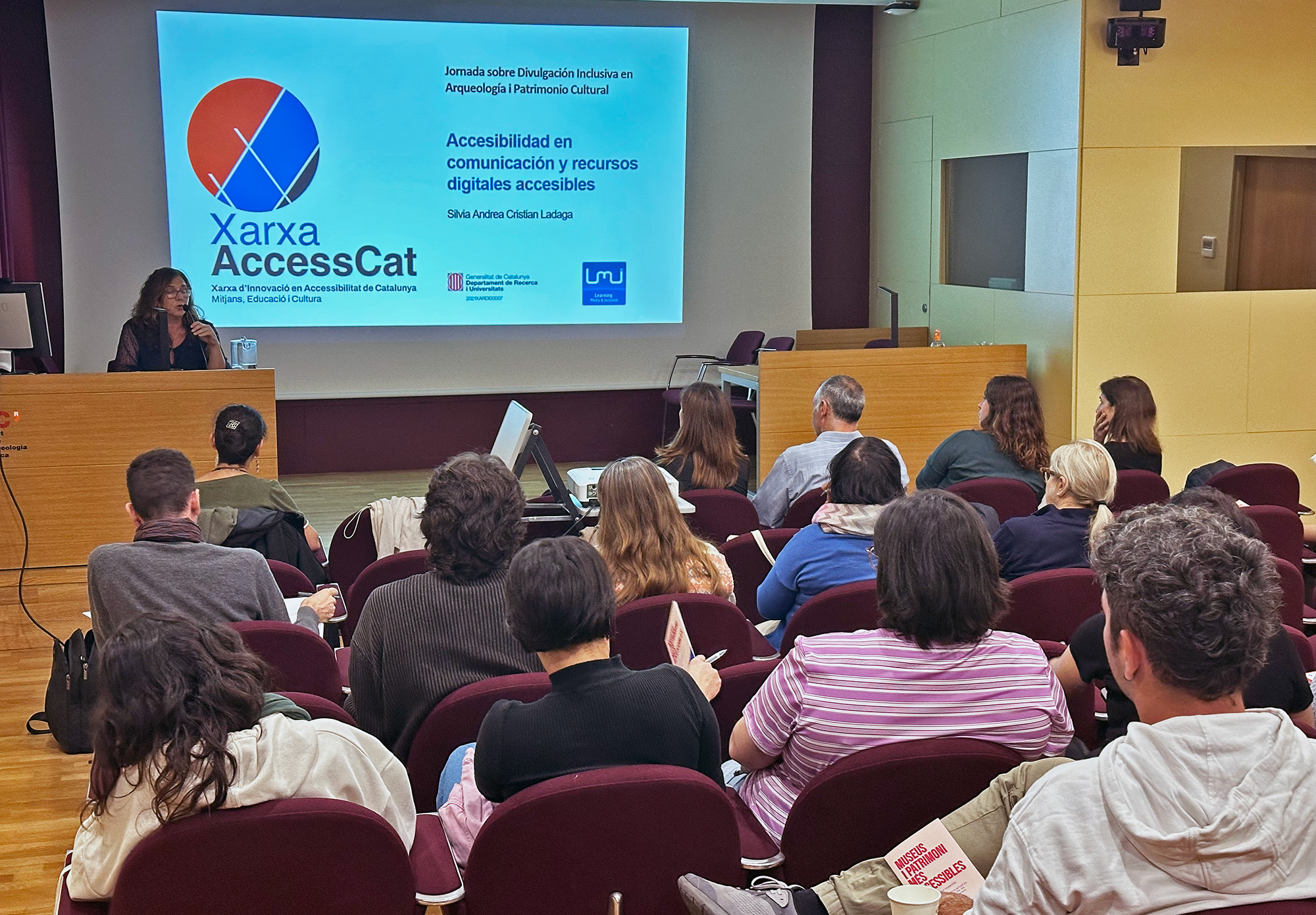 La investigadora Cris Ladaga, de LMI, impartint una xerrada sobre accessibilitat. De fons, una presentació amb el logo de la xarxa.