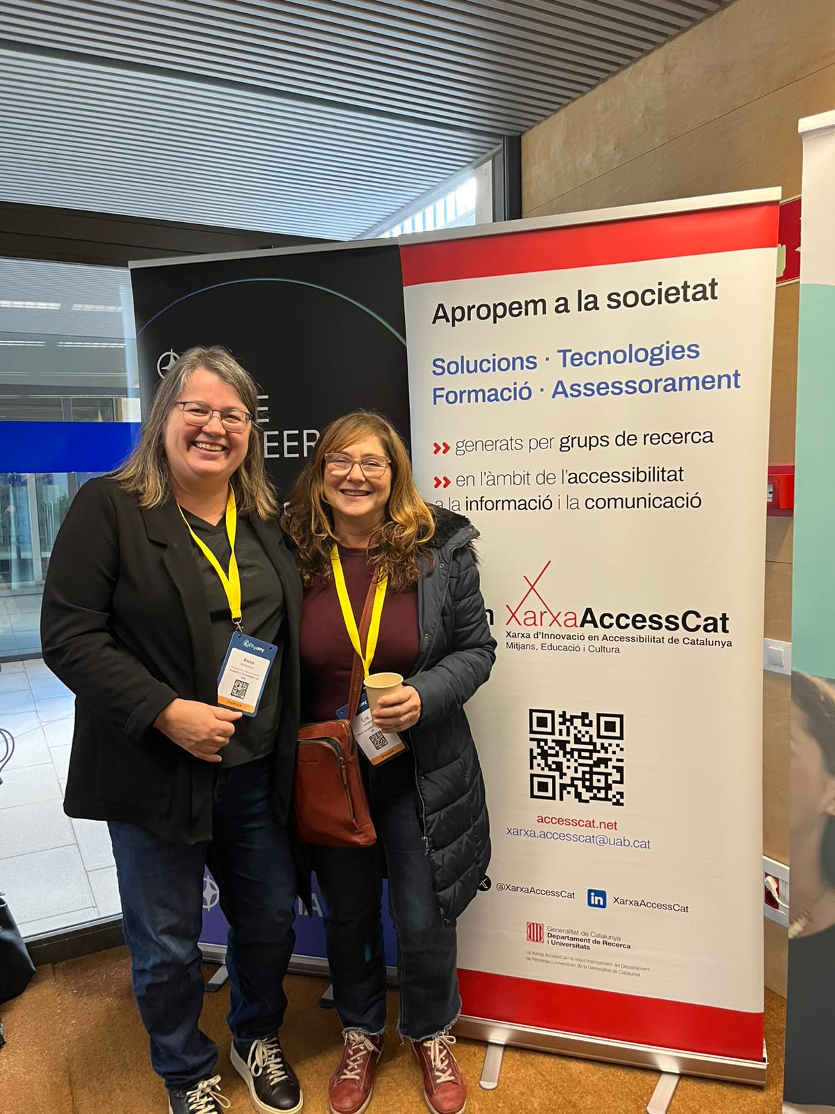 Researchers Anna Matamala and Cris Ladaga at a11yConf, in front of a network roll-up. The roll-up reads: “We bring society solutions, technologies, training, and guidance generated by research groups in the field of information and communication accessibility.”