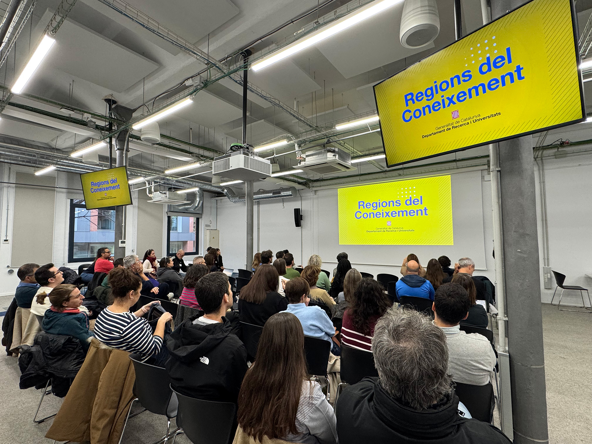 Unes 40 persones assistents a l'esdeveniment miren unes pantalles grogues amb el text "Regions del coneixement" escrit.